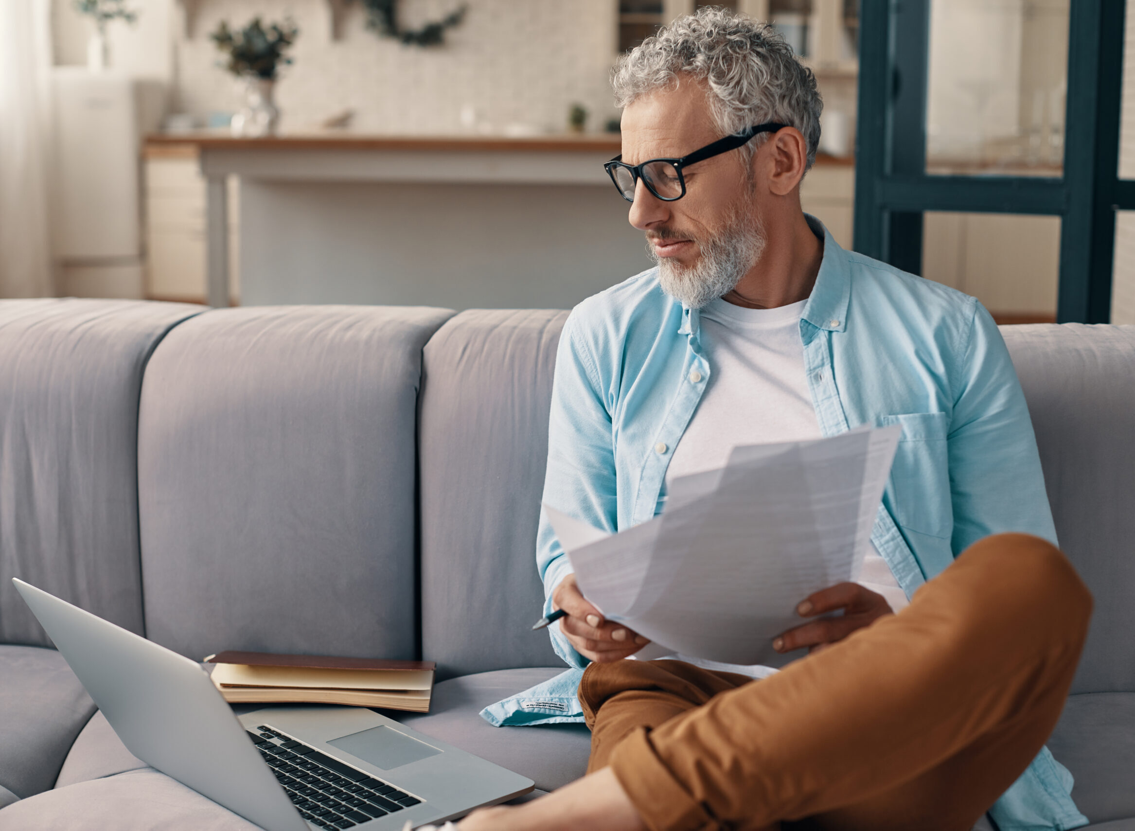 A senior man filling in a driving licence application form while sitting on the sofa at home with a laptop next to him