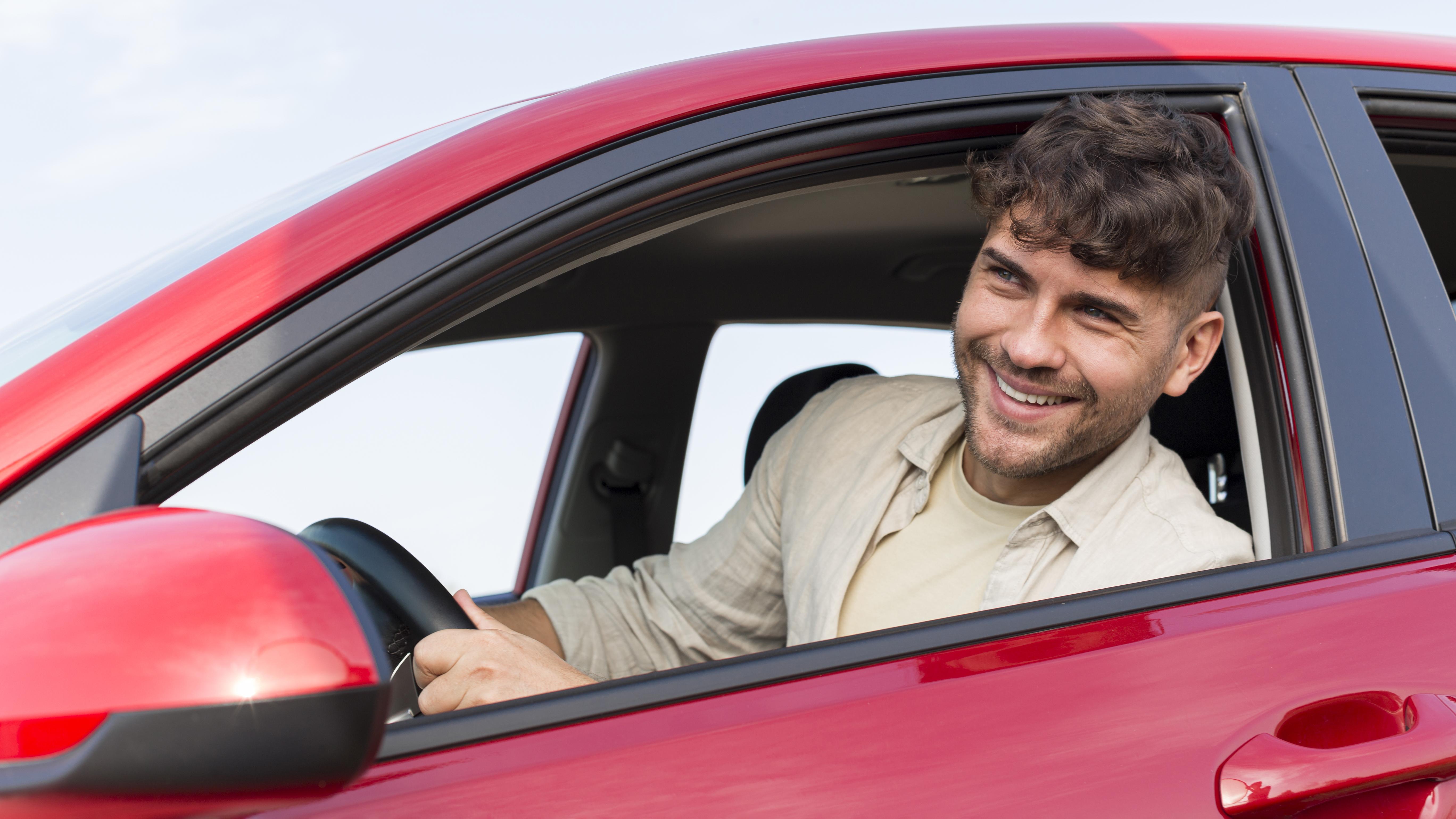 A young man with a wide smile driving a car, holding the steering wheel, and looking at the scenery through the windshield.